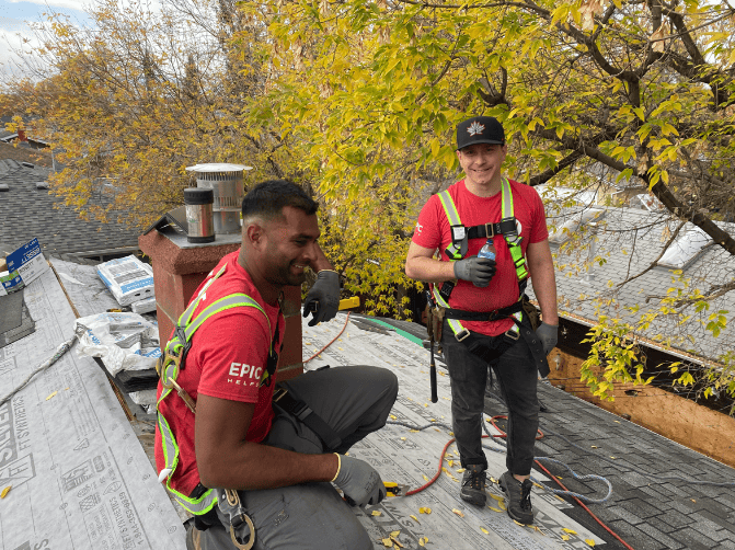 Two roofers with tools on a partially installed roof with fall trees in the background.