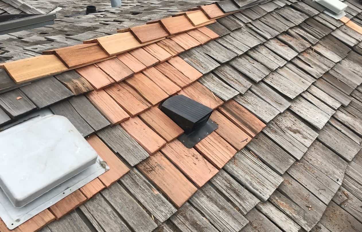 Close-up of a weathered shingle roof with a vent and a mix of gray and terracotta-colored tiles.