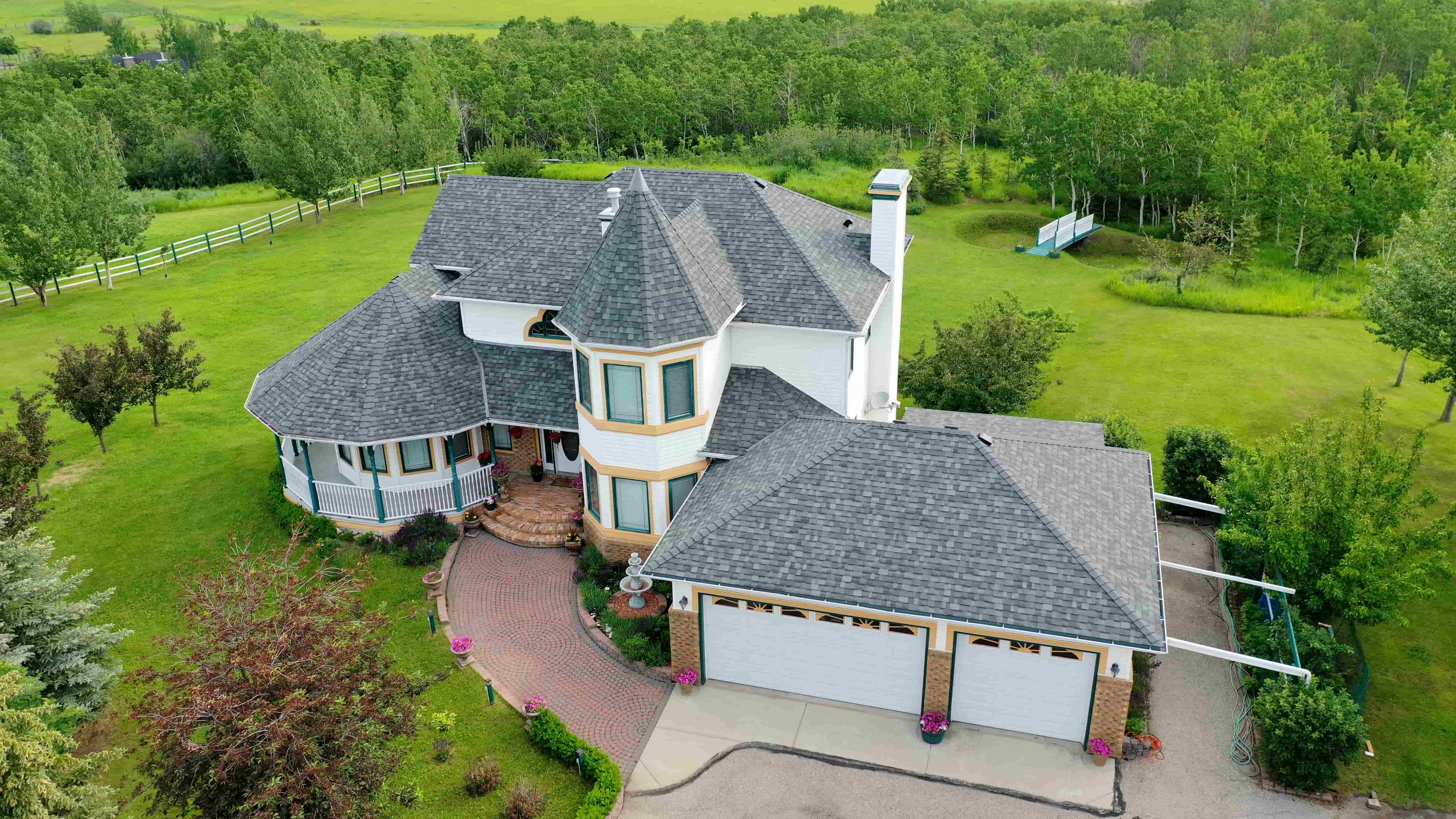 Aerial view of a large white home with grey shingles, a turret, and a big green yard surrounded by trees.