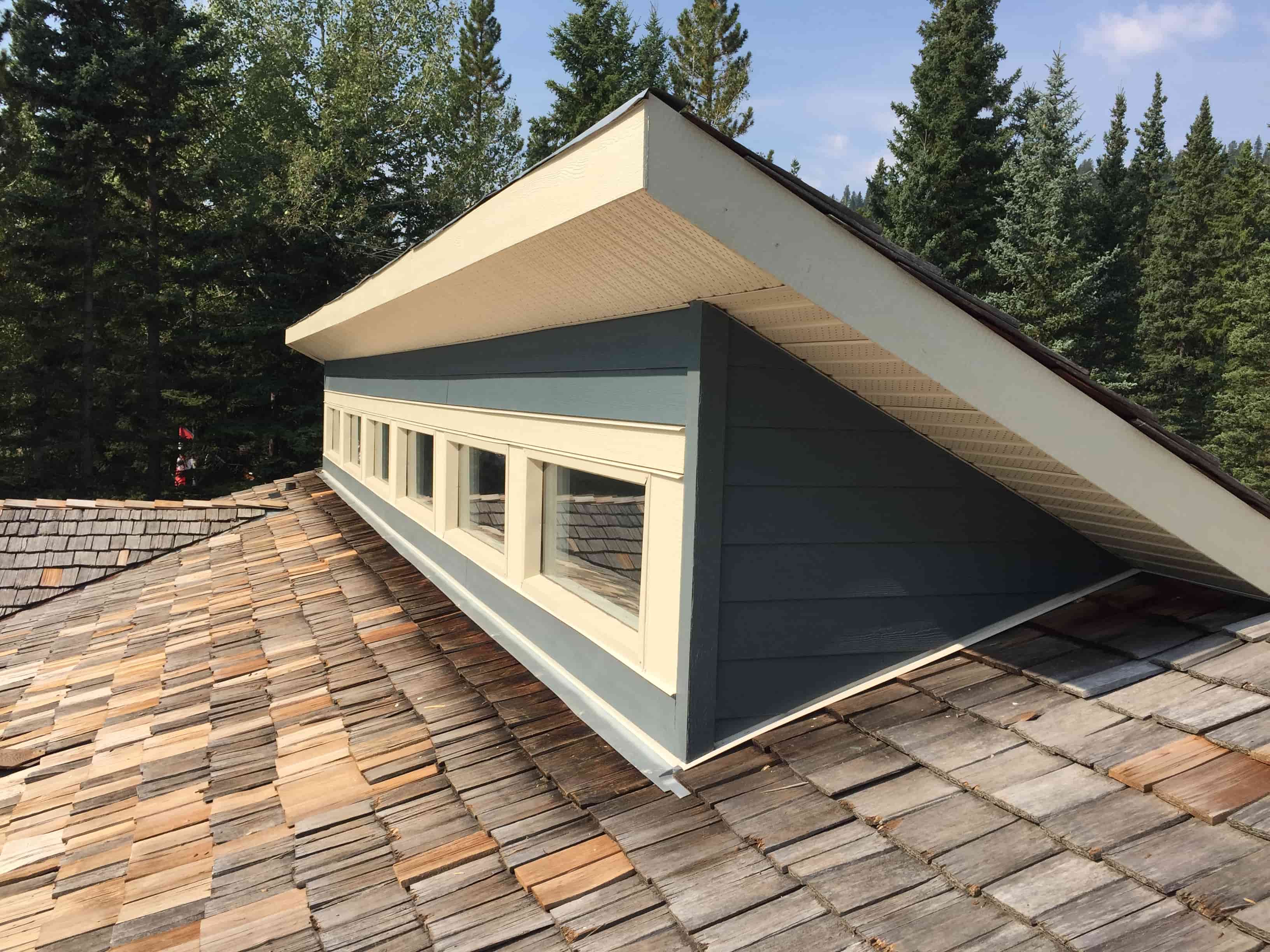 Wooden cedar shake roof built around a blue dormer with a row of small windows, surrounded by pine trees.