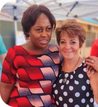 Two smiling women embracing, one wearing a geometric-patterned dress and the other in a polka-dot top, at an outdoor social event