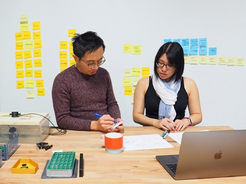 Two designers standing at a workbench, discussing and reviewing a set of sketches and prototypes.