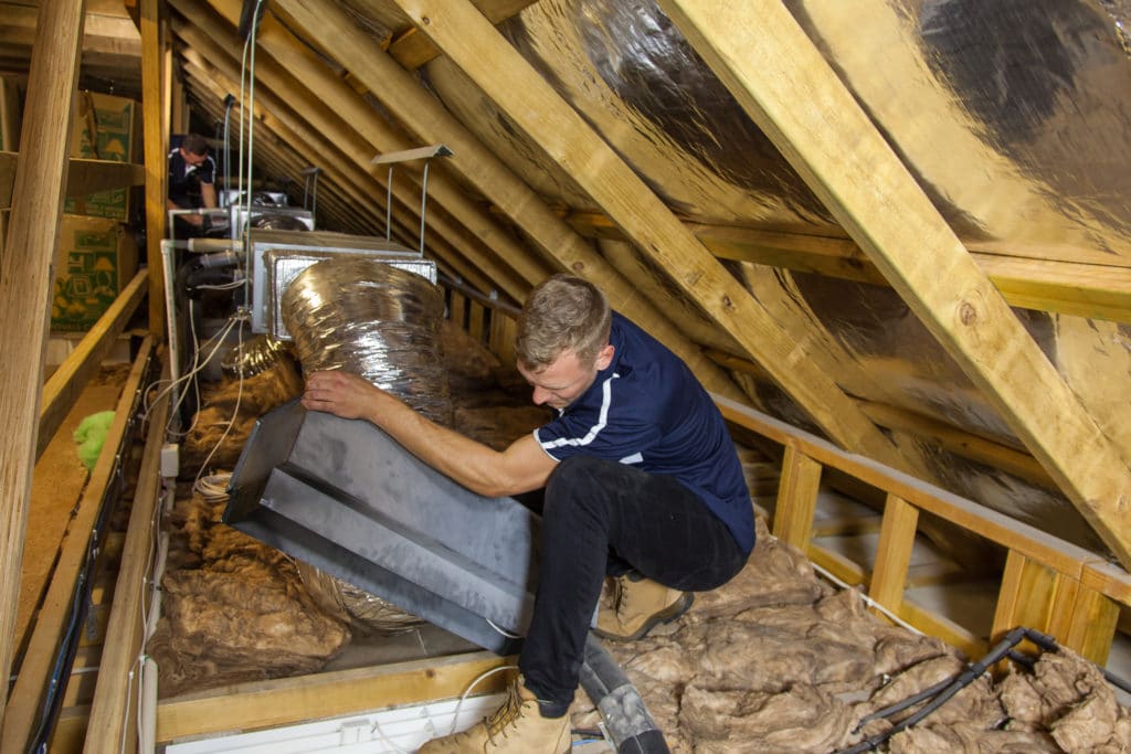 Residential HVAC technician fitting an insulated duct to a metal plenum inside a roof cavity during ducted air conditioning installation