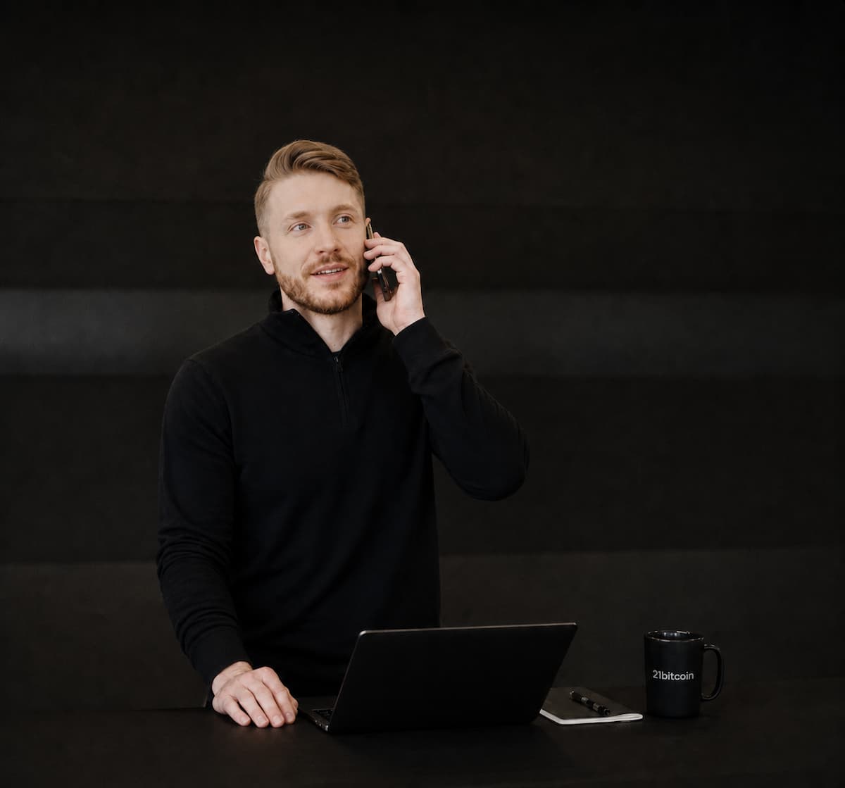 Mann telefoniert neben Laptop, Notizblock und Kaffeetasse mit 21bitcoin-Logo auf schwarzem Tisch.