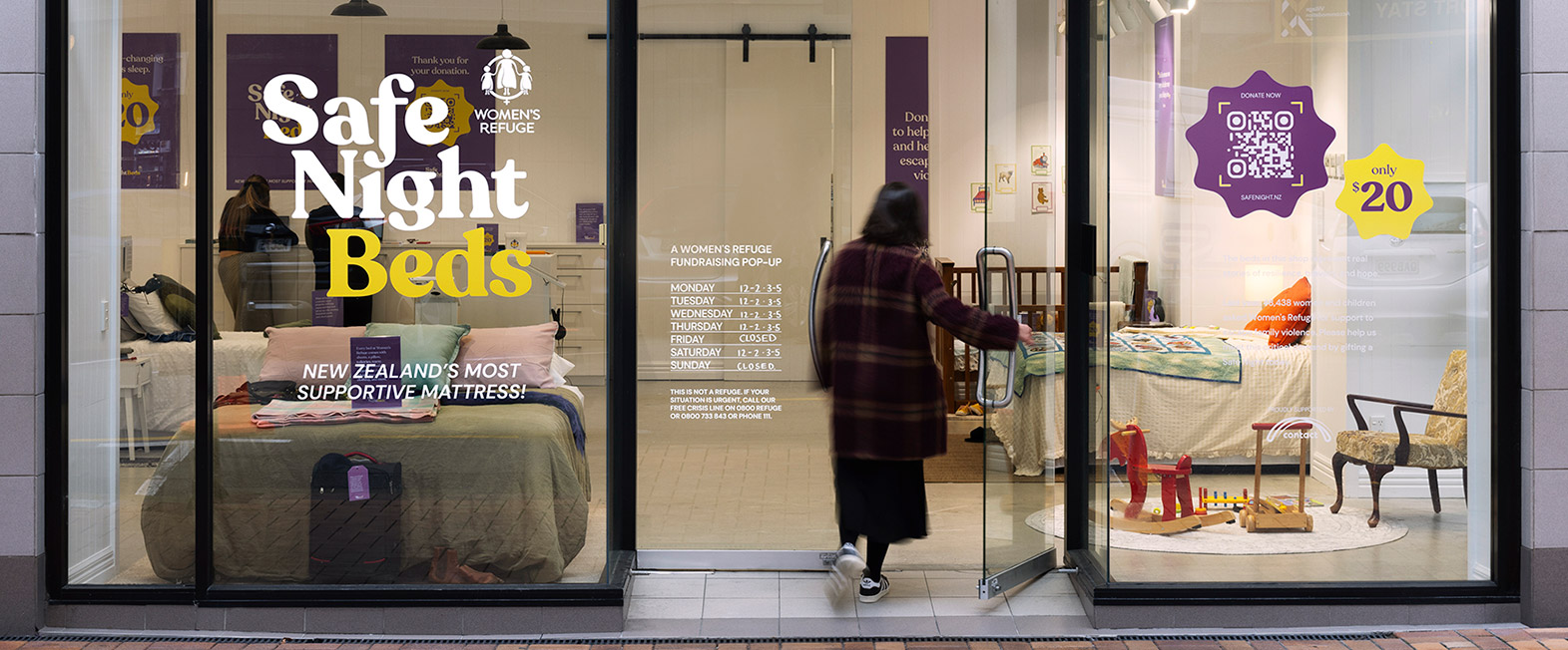 A woman enters a glass-fronted pop-up shop displaying Safe Night Beds fundraiser for Women's Refuge, showing beds and children's toys inside.