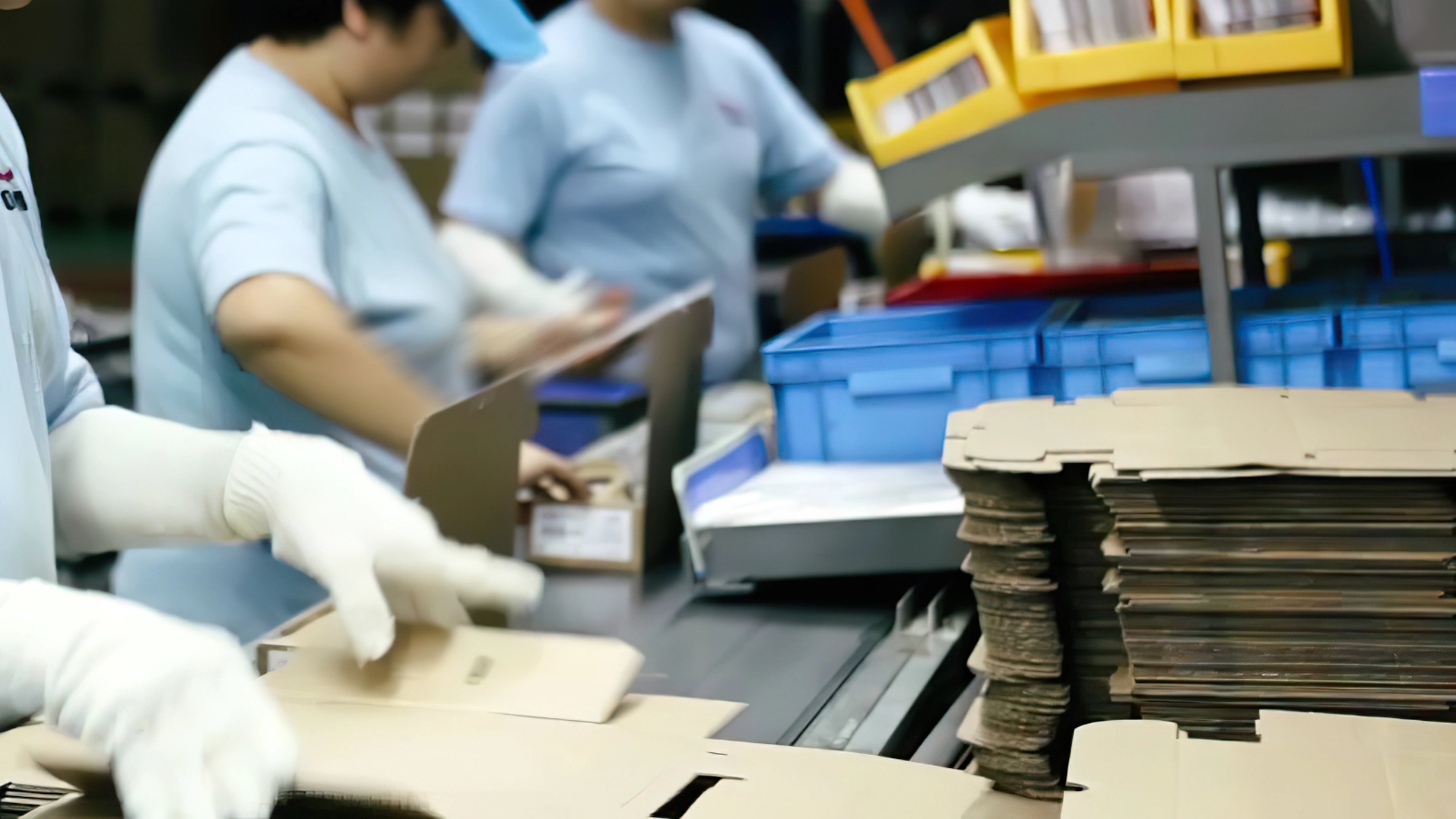 Workers assemble and fold cardboard boxes on a factory conveyor belt wearing white gloves.