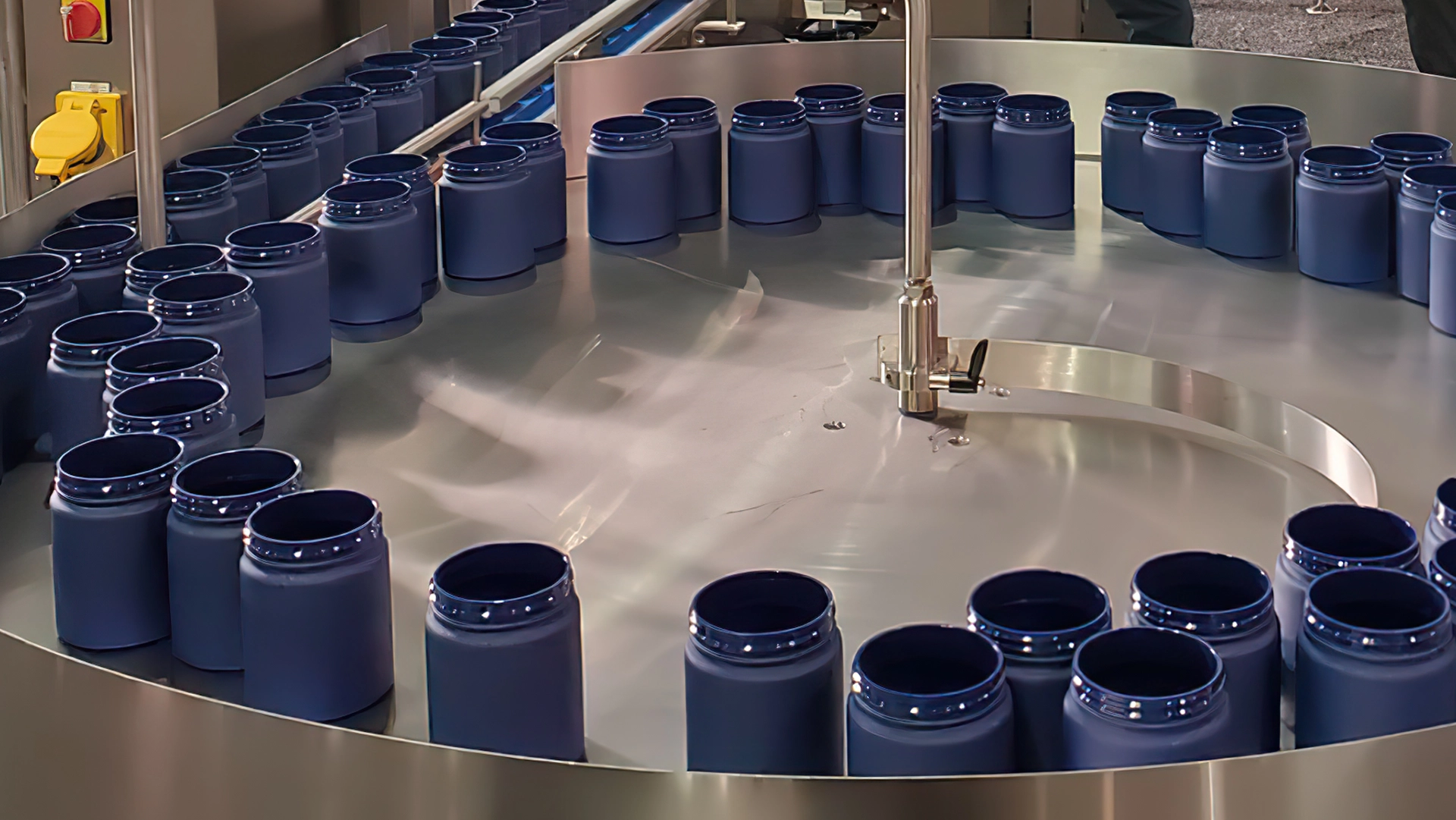 Circular assembly line with multiple empty blue plastic jars arranged around a rotating metal platform in a manufacturing facility.