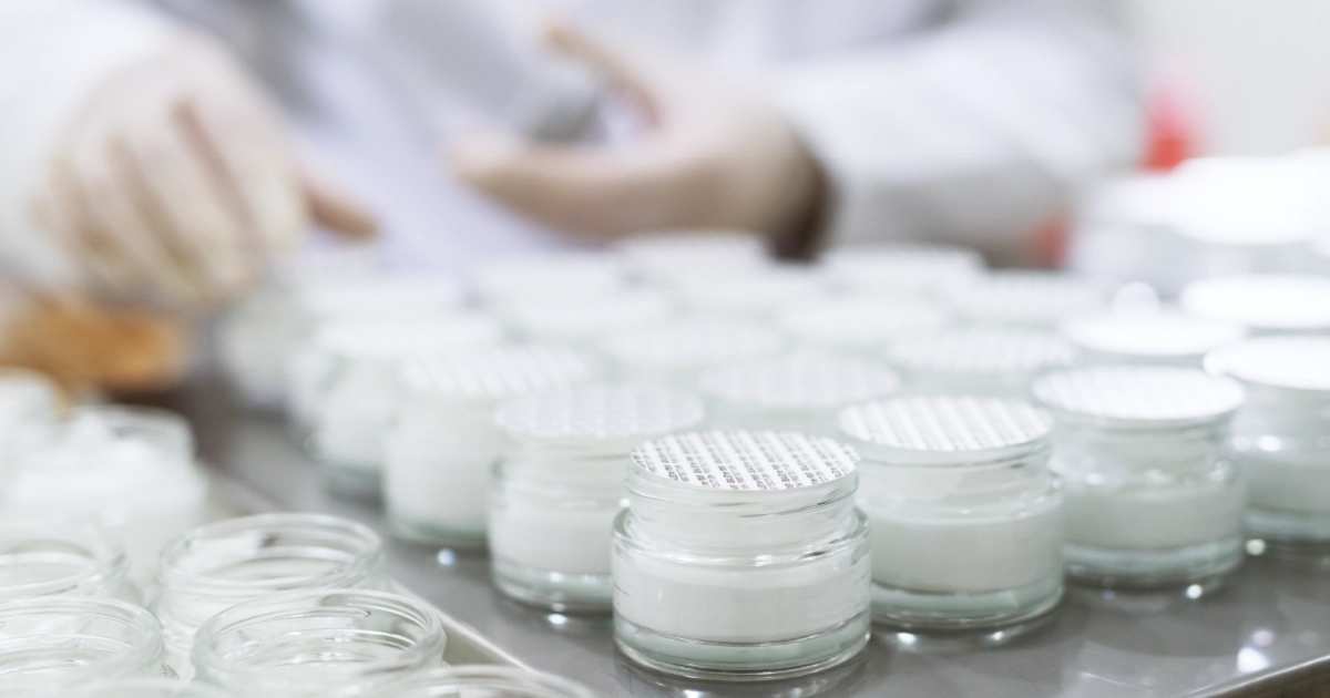 Rows of small glass jars filled with white cream on a production line with a person wearing gloves handling them in the background.