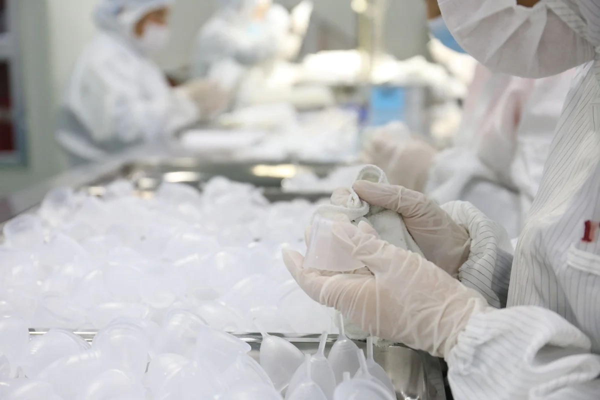 Worker wearing gloves and protective gear inspecting and polishing translucent menstrual cups in a manufacturing facility.