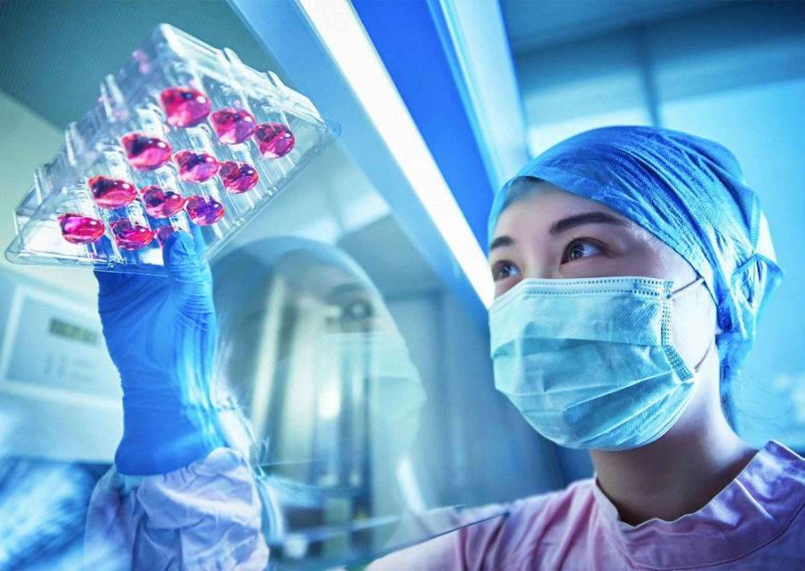 Scientist wearing mask and blue hair covering inspecting a tray with pink liquid-filled vials in a laboratory.
