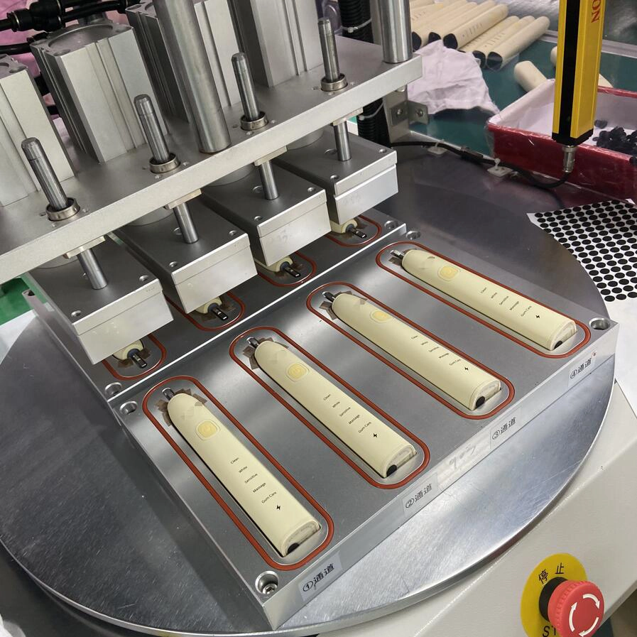 Four white electric toothbrush handles positioned in a metal fixture on a manufacturing assembly line.