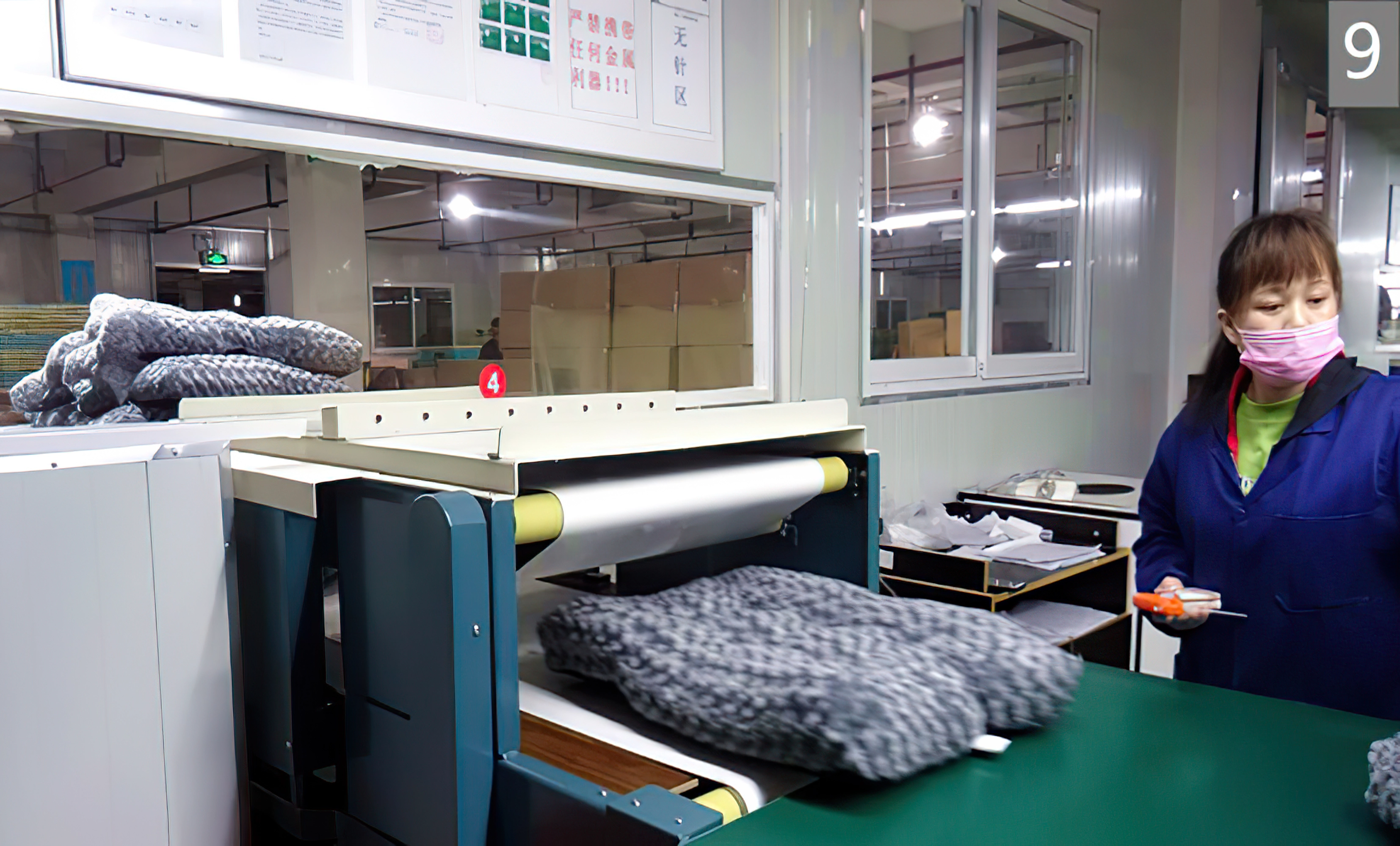Worker in a face mask operating machinery that processes folded fabric pieces in a factory setting.