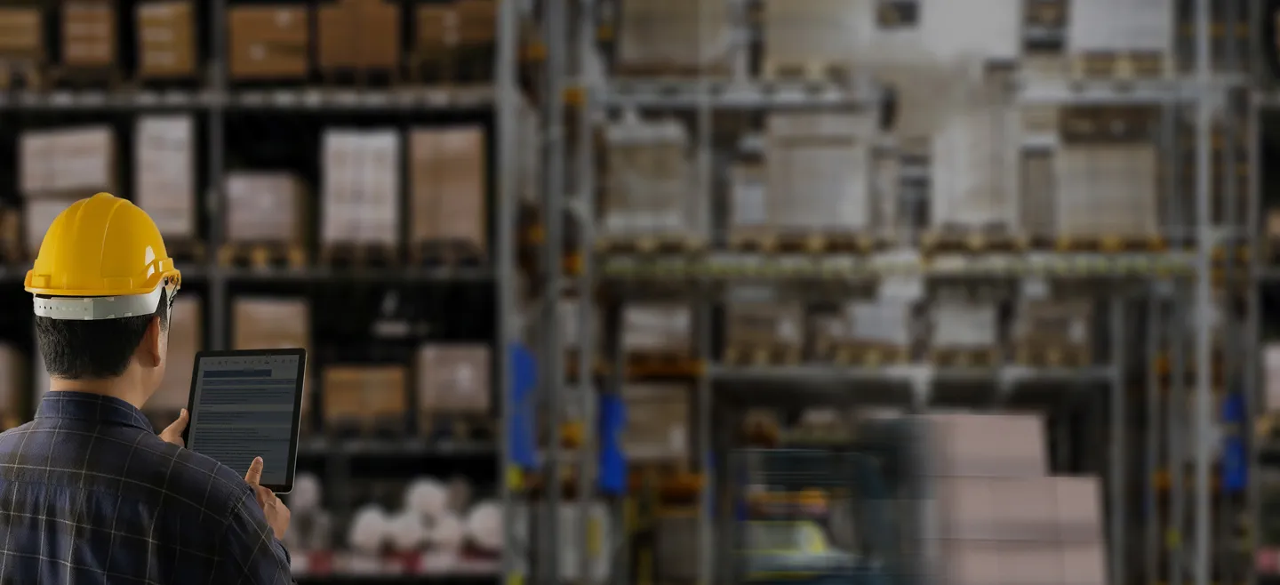 Worker wearing a yellow hard hat using a tablet inside a warehouse with shelves full of boxes.