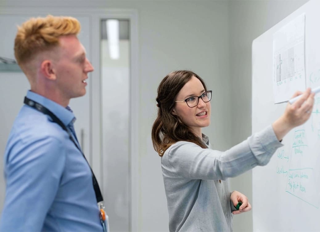 Woman pointing at a whiteboard with charts and notes while a man watches attentively in an office setting.
