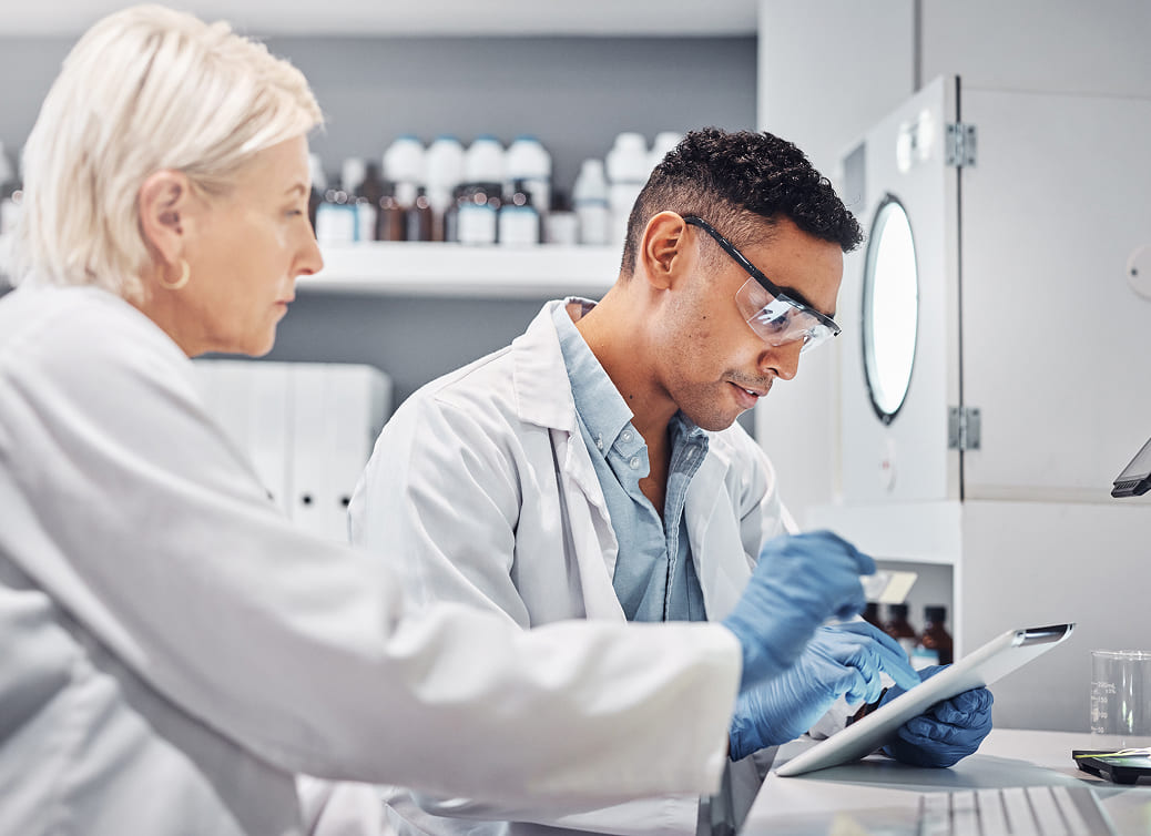Two scientists in lab coats working together in a laboratory, one using a tablet and wearing safety glasses and gloves.