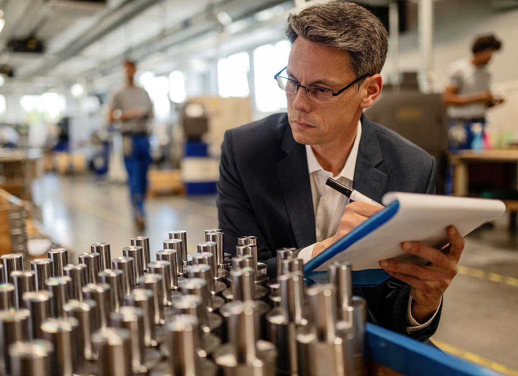 Man in glasses and suit inspecting and taking notes on metal cylindrical parts in an industrial setting.
