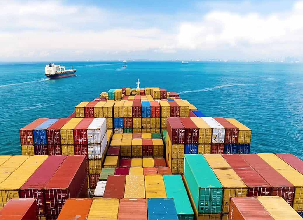 View from a cargo ship deck showing stacked colorful shipping containers with another ship and calm blue sea in the background.