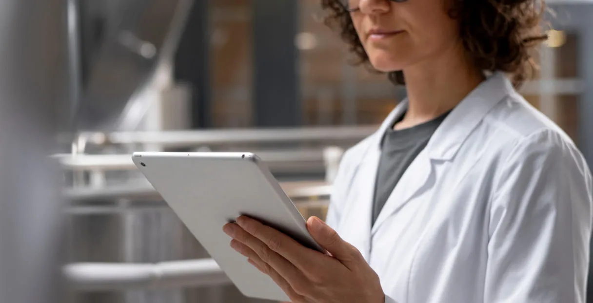 Woman wearing a white lab coat holding and looking at a tablet device indoors.