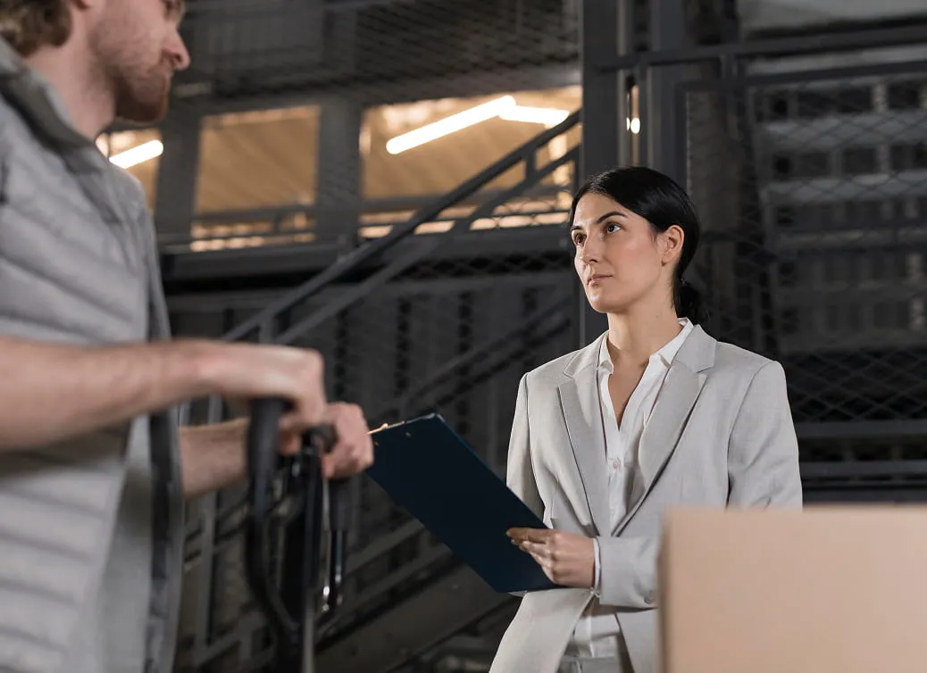 Woman in a light gray suit holding a clipboard and talking to a man operating a hand truck in an industrial or warehouse setting.