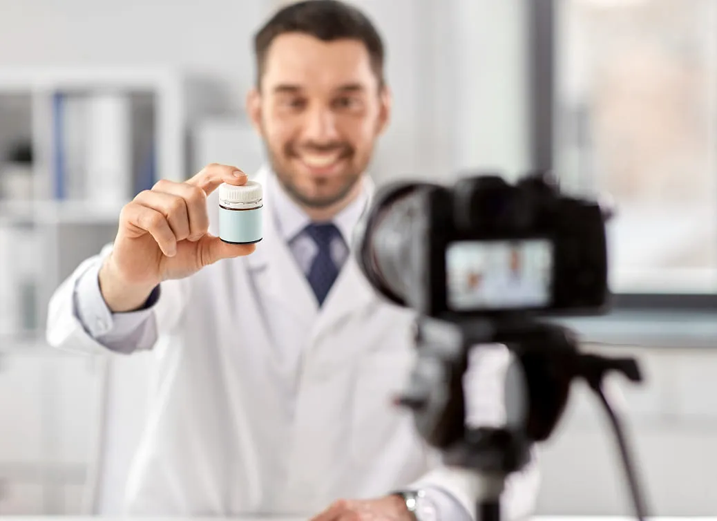 Smiling male doctor in white coat holding a small medicine bottle while recording on a camera in a bright office.