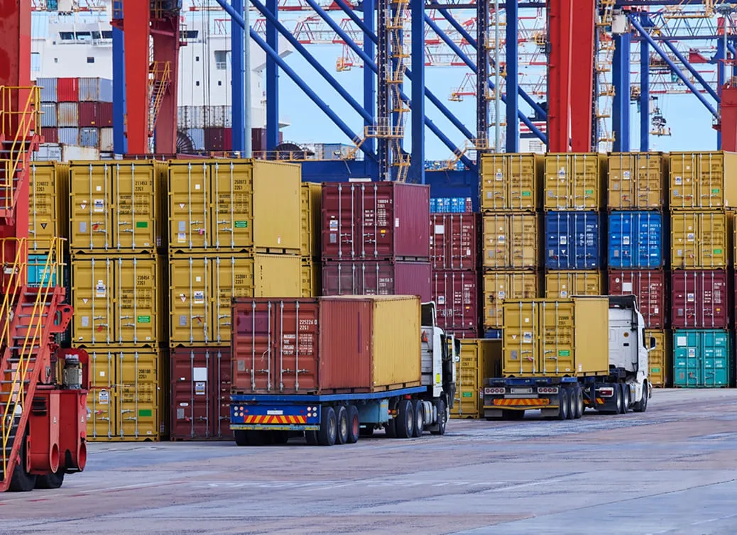 Two trucks transporting stacked yellow and red shipping containers at a busy port with cranes in the background.