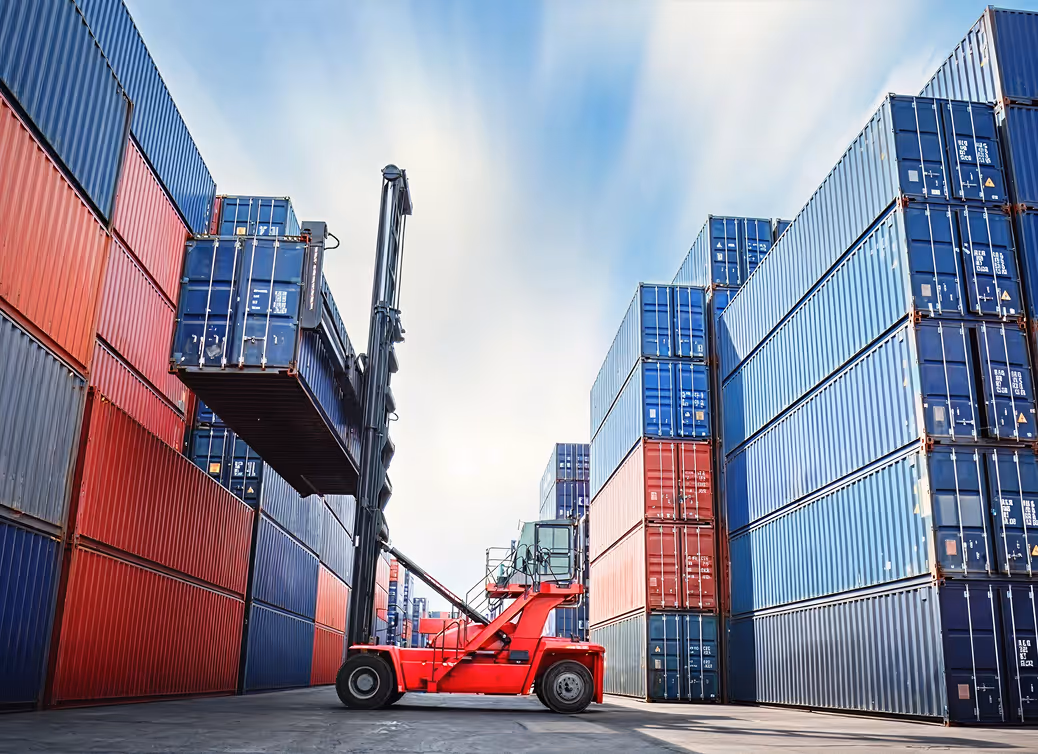 Red forklift lifting a blue shipping container between tall stacks of red and blue containers under a partly cloudy sky.