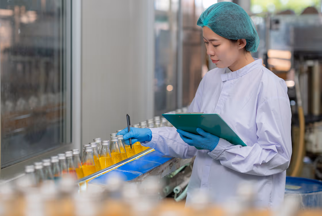 Woman in protective clothing and hairnet inspecting and recording data on bottles of yellow liquid in a production line.