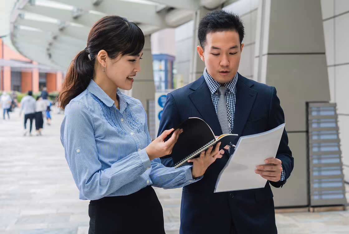 Two young professionals outdoors reviewing documents together, one holding a notebook and the other a paper.