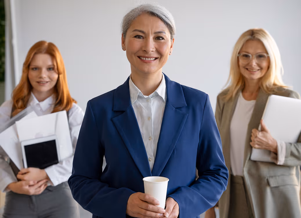 Smiling professional woman in blue blazer holding a paper cup, flanked by two women carrying office documents and a laptop.