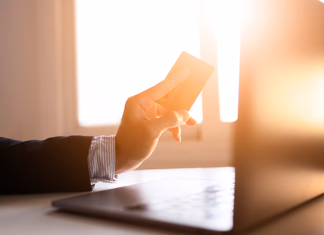 Person in business attire holding a credit card above a laptop with sunlight streaming through a window.