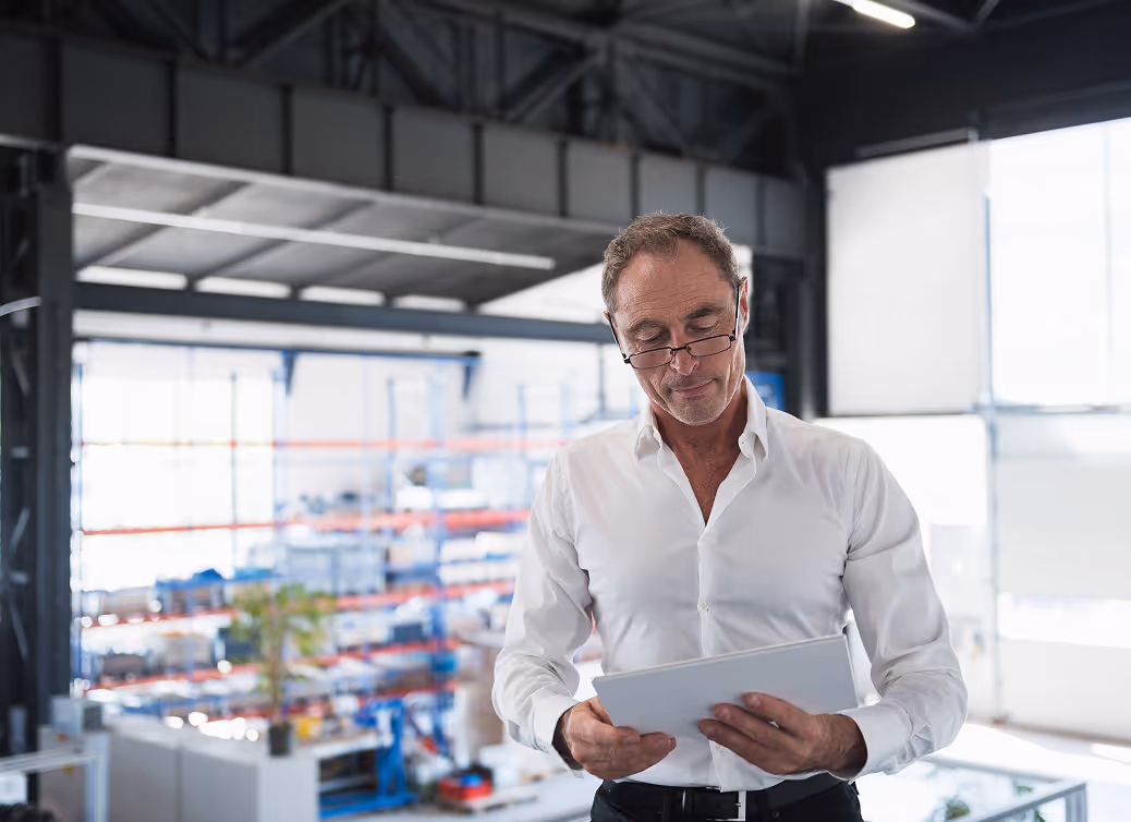 Middle-aged man wearing glasses looking at a tablet in a bright modern warehouse.