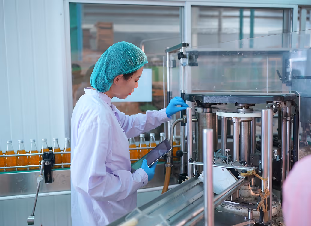 Woman in protective clothing and hairnet operating machinery on a beverage bottling line while holding a tablet.