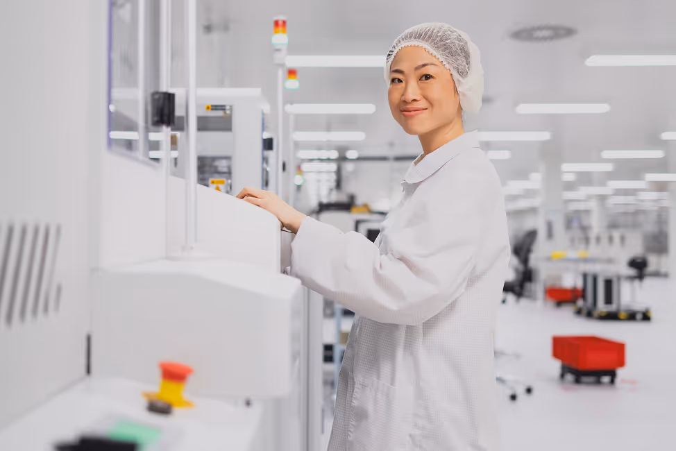 Smiling female technician in cleanroom attire operating a machine in a bright, modern laboratory.