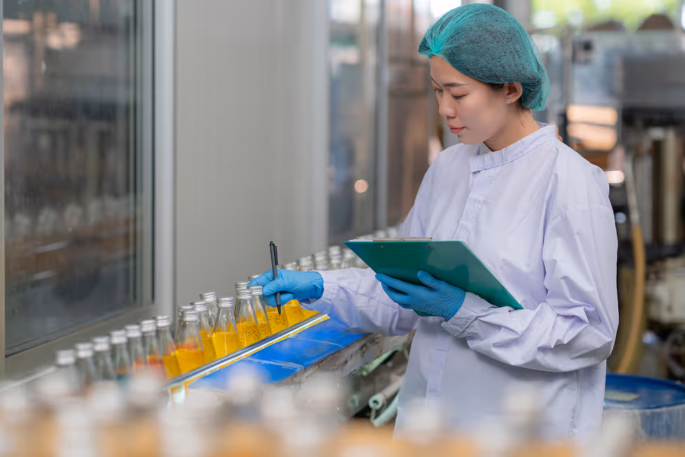 Female quality control technician in protective clothing inspecting and marking bottles of orange liquid on a production line.