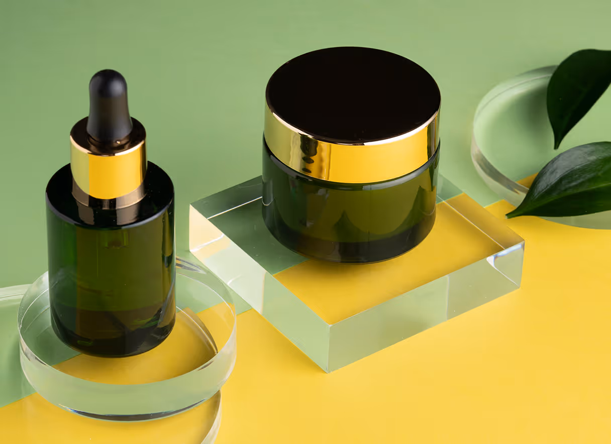 Two dark green cosmetic containers with gold lids on transparent stands against a green and yellow background, with a green leaf on the right.