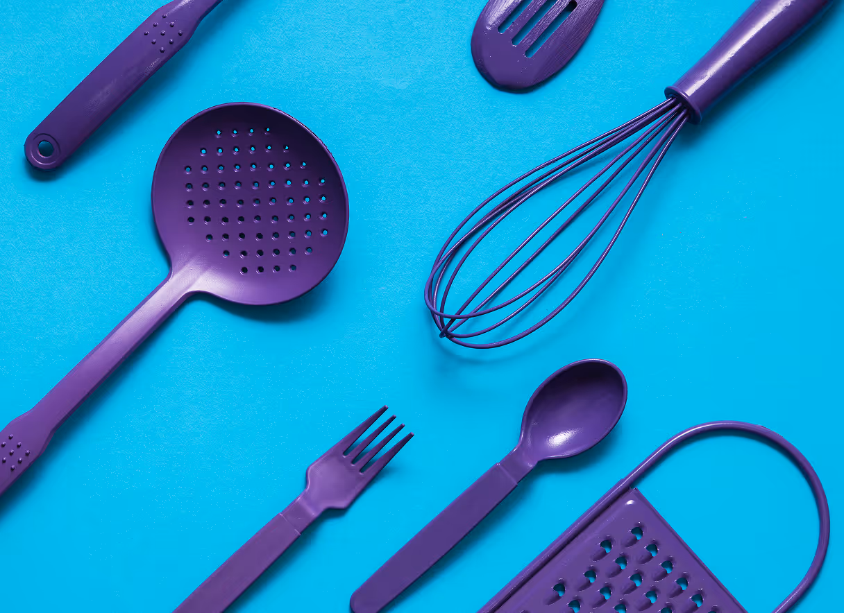 Flat lay of assorted purple kitchen utensils including a whisk, fork, spoon, slotted spoon, grater, and spatula on a bright blue background.