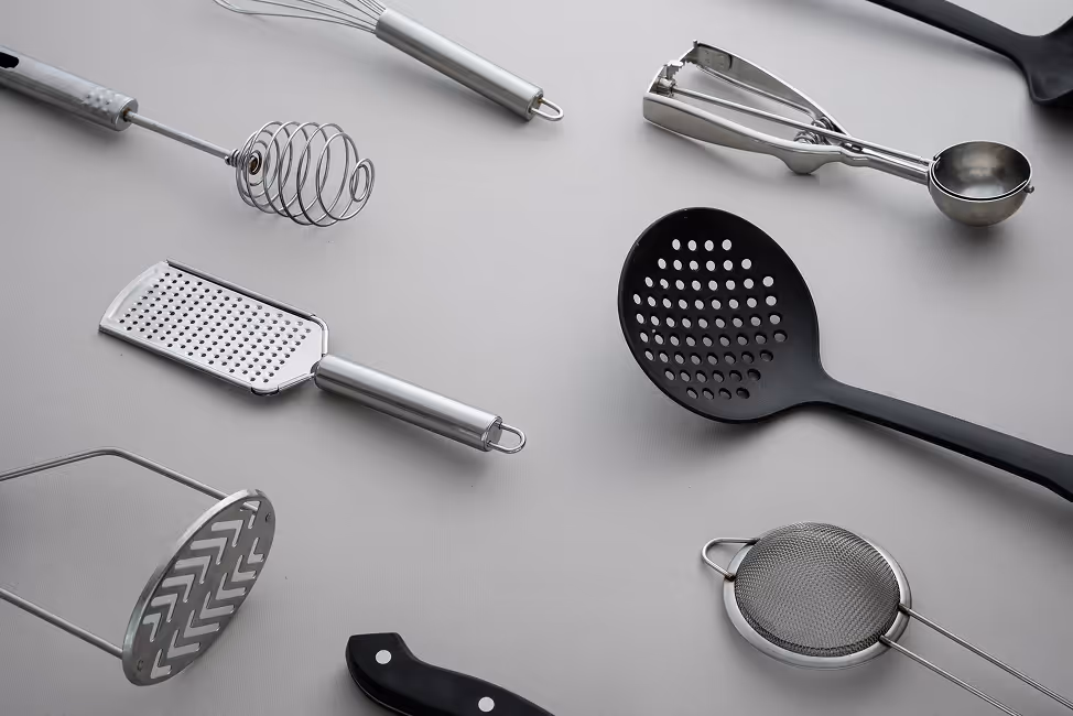 Various kitchen utensils including a whisk, grater, potato masher, slotted spoon, ice cream scoop, and strainer arranged on a light gray surface.