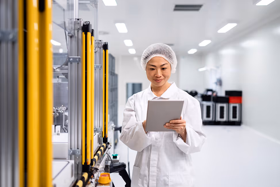 Person in white lab coat and hairnet holding a tablet, standing next to industrial machinery in a clean, well-lit facility.