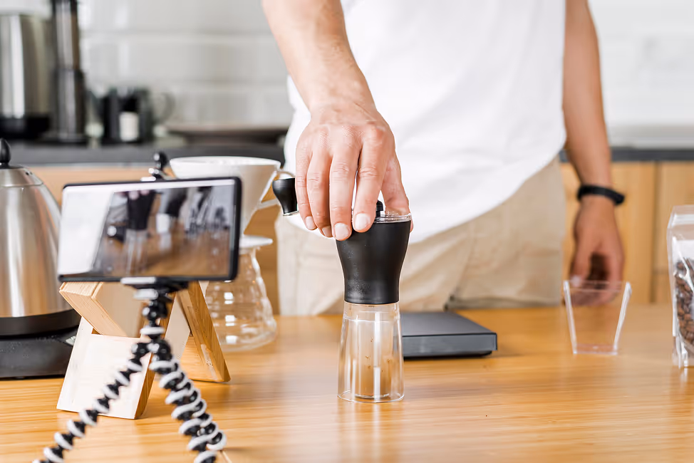 Person grinding coffee beans with a hand grinder on a wooden table, with a smartphone on a tripod recording.