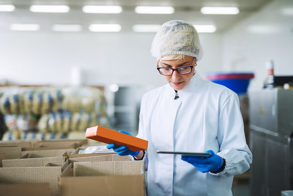 Woman in protective clothing and hairnet inspecting an orange package with a tablet in a packaging facility.