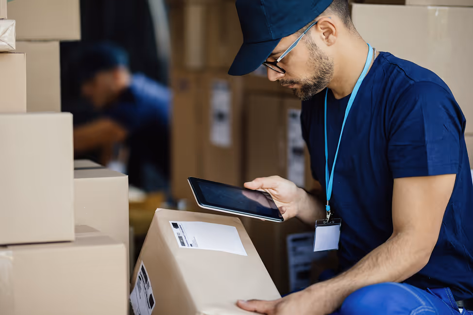 Warehouse worker wearing a cap and badge scanning a package label with a handheld tablet.