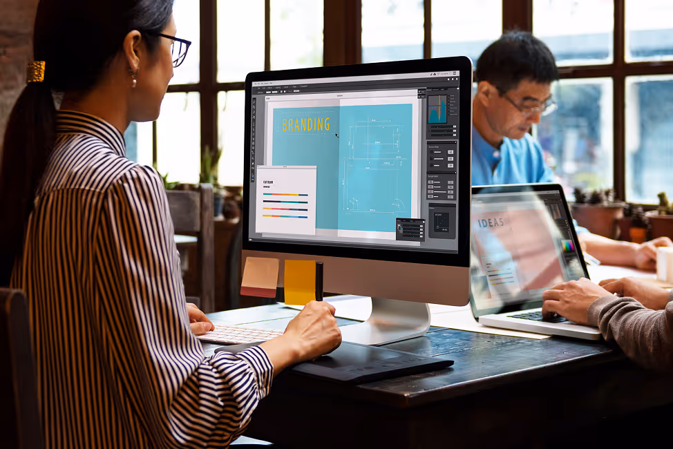 Woman working on desktop computer with branding design open, man in background using laptop.