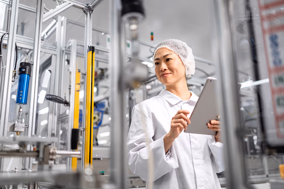 Laboratory or cleanroom technician in white protective wear and hairnet using a tablet amid industrial equipment.
