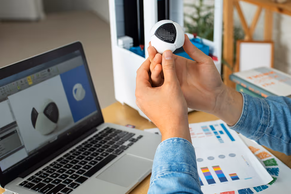 Person holding a small white spherical object with a black triangular section, next to a laptop displaying a 3D model of the object and papers with colorful charts on a desk.