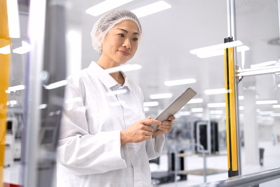 A woman in a white lab coat and hairnet using a tablet in a clean, modern laboratory or manufacturing facility.