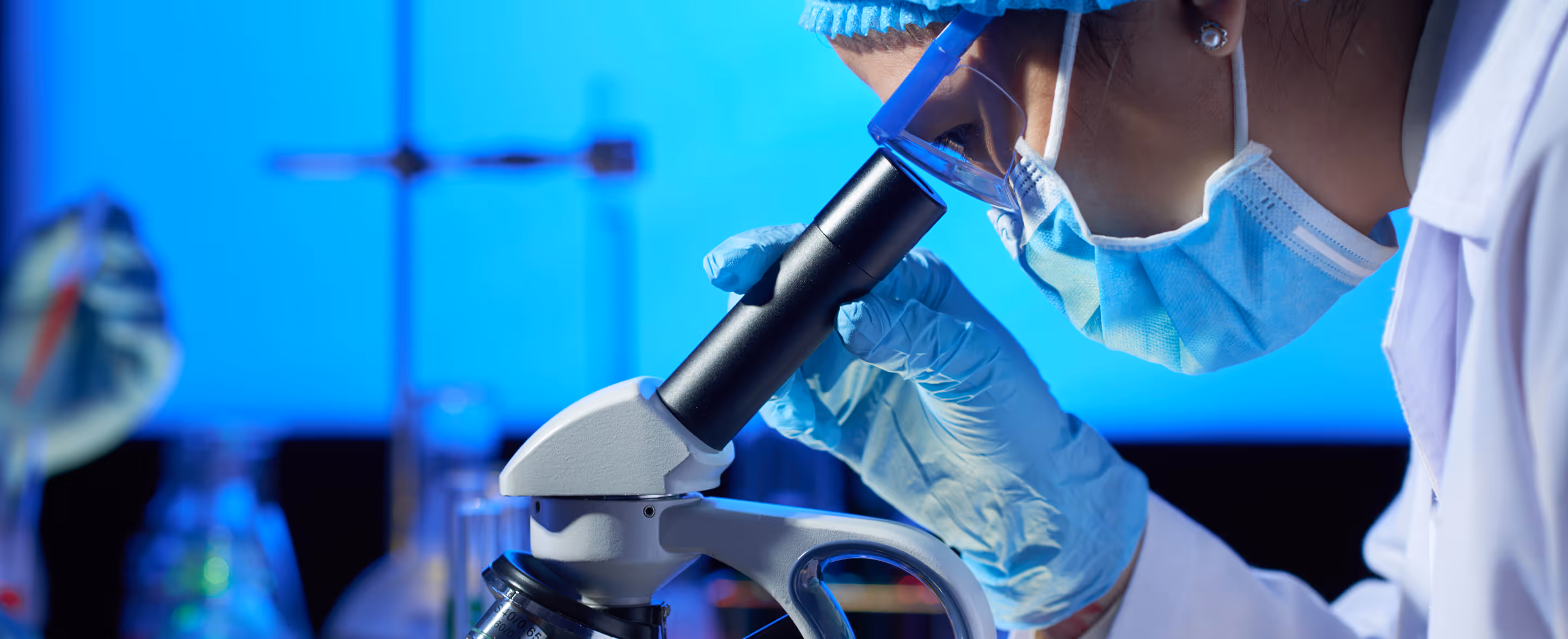 Scientist wearing a mask, gloves, and protective eyewear looking through a microscope in a laboratory.