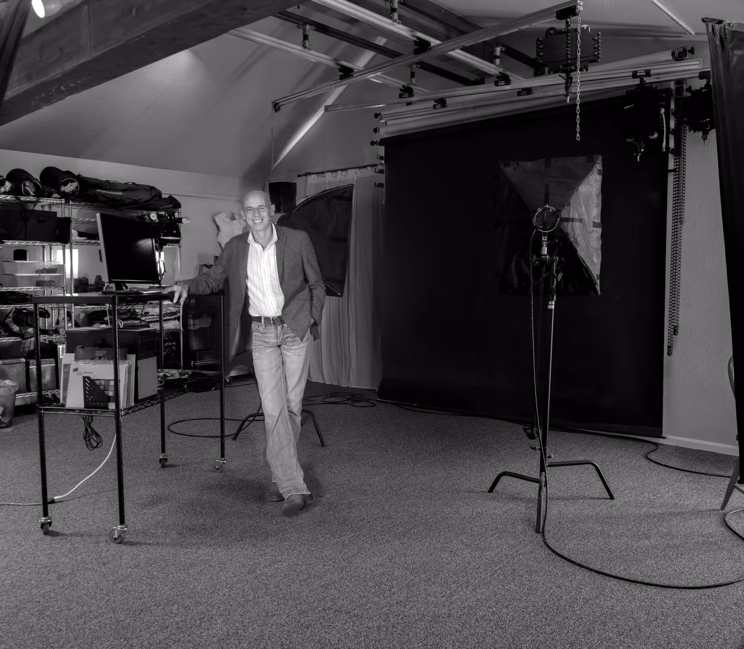 Smiling man in jeans and blazer leaning on a table in a photography studio with lighting equipment and backdrop.