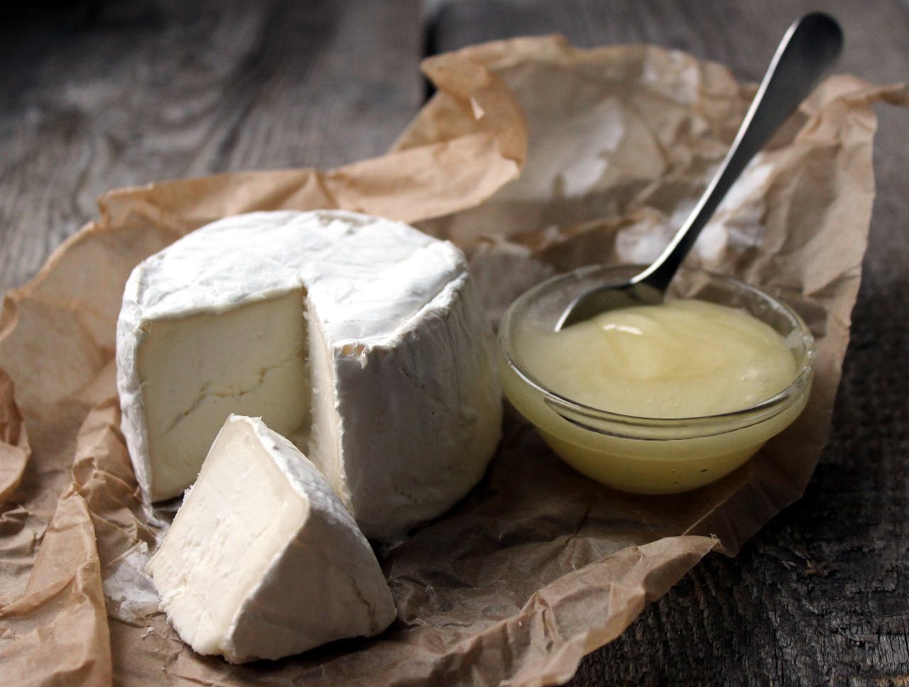 A round cheese wheel with a cut piece sits on crumpled paper, accompanied by a small bowl of creamy sauce and a spoon.