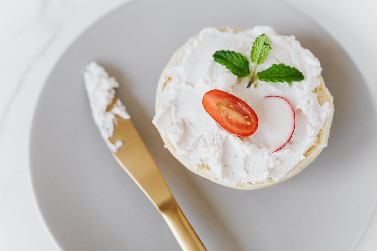 A round bread topped with creamy spread, a slice of tomato, a radish, and a mint leaf, accompanied by a gold knife on a gray plate.