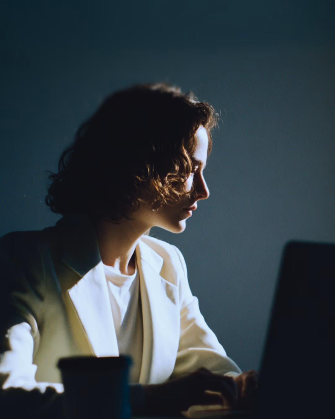 Profile of person with curly hair working on laptop in low light wearing a light-colored blazer.